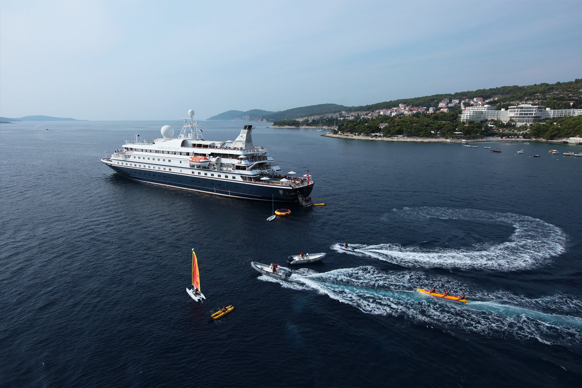 A large expedition cruise ship with distinctive dark hull and white superstructure is anchored in crystal-clear turquoise waters off a scenic Mediterranean coastal town. Small water sports boats and kayaks are visible in the foreground, while a picturesque hillside village with white buildings and lush green vegetation rises behind the vessel.