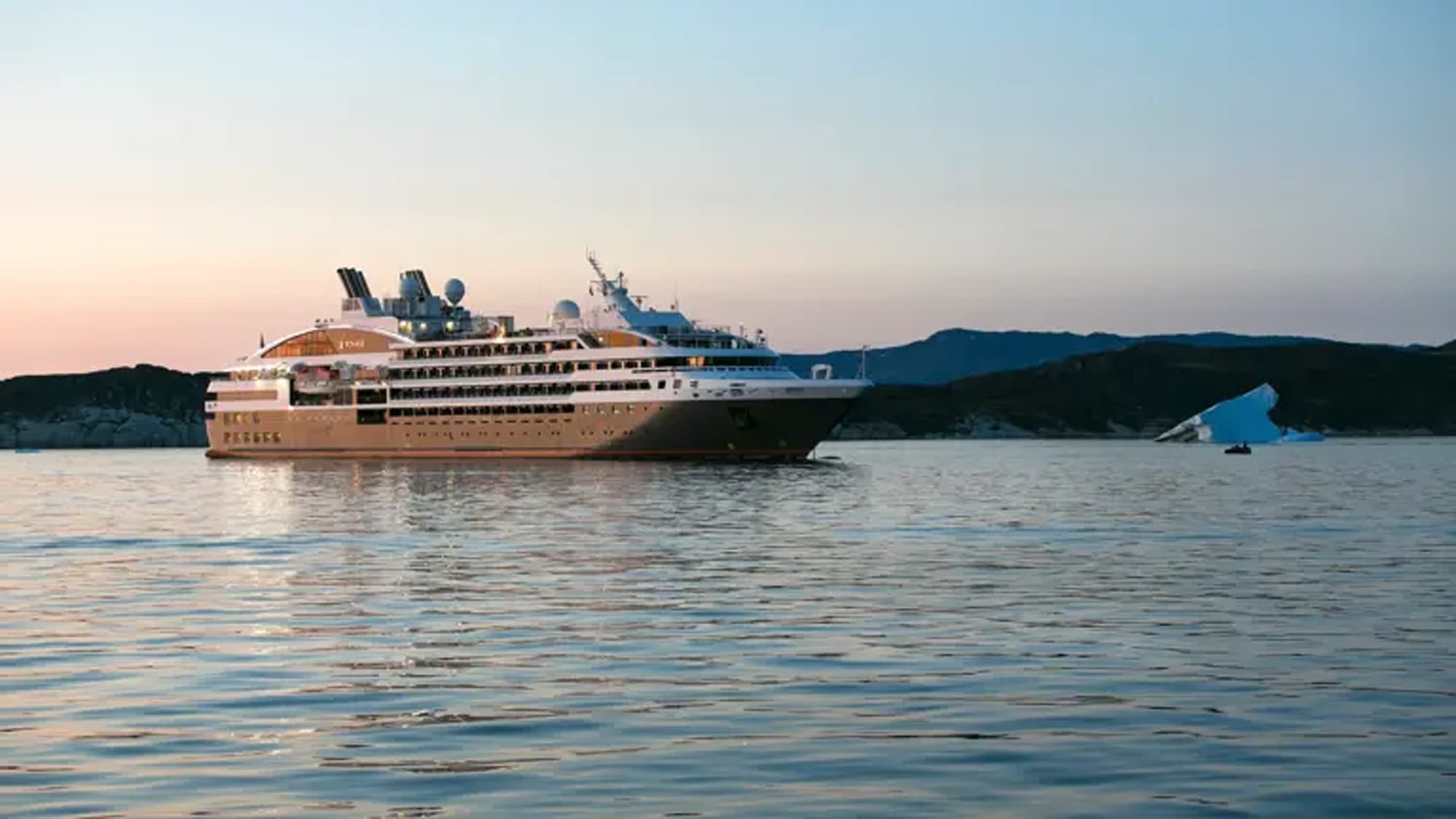 Luxury Cruise Ship in Arctic Waters A modern expedition cruise ship anchors in calm Arctic waters near Bisko Bay, with snow-capped mountains and an iceberg visible in the background during twilight hours. The vessel features a distinctive design with multiple decks and contemporary architecture typical of luxury expedition cruising.