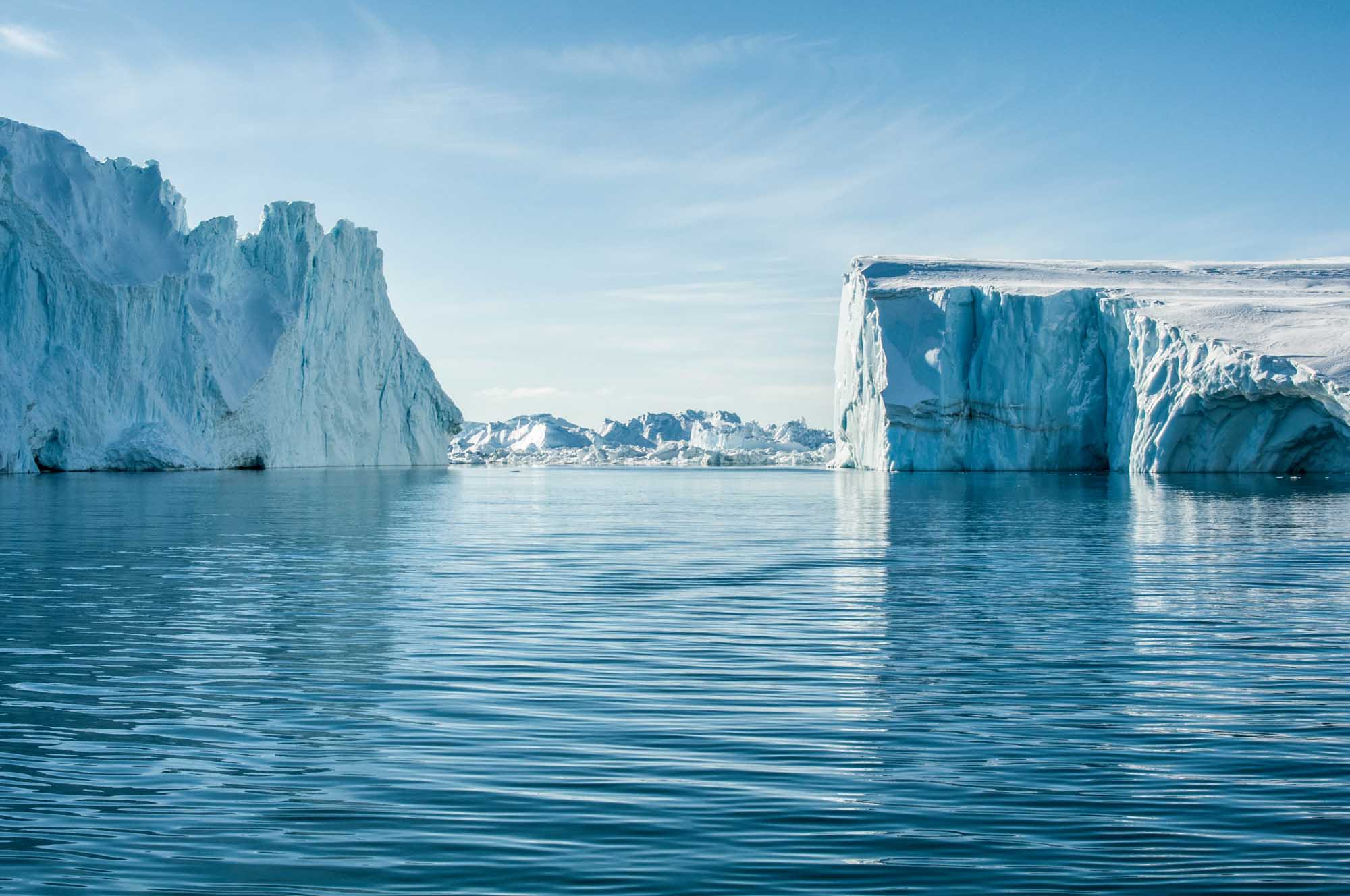 Towering white and blue icebergs float in calm turquoise Arctic waters, with a small Greenlandic