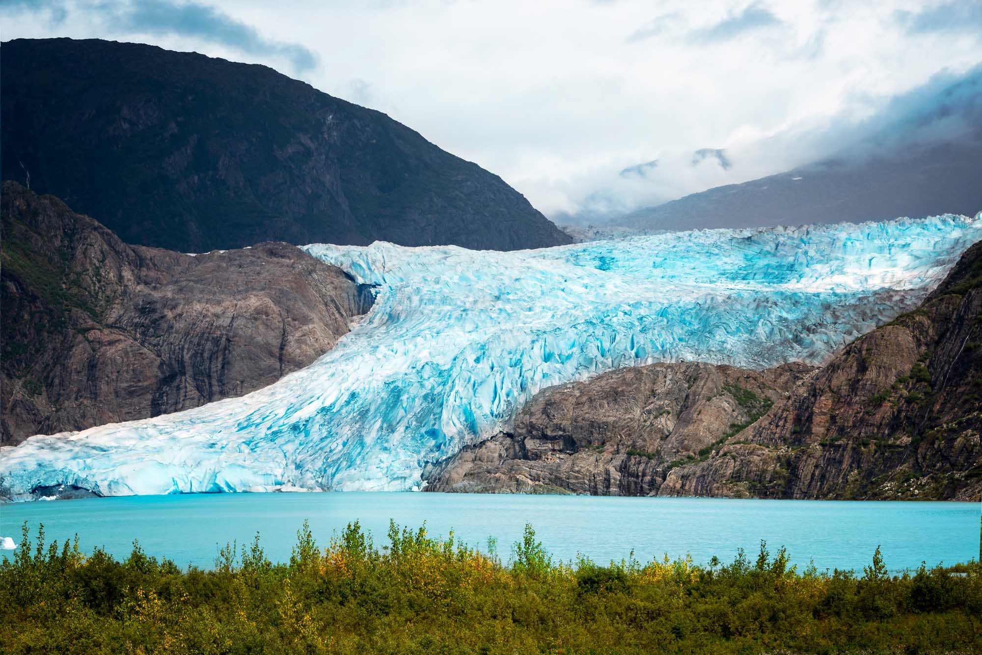 A stunning turquoise glacier cascades into a pristine glacial lake, surrounded by dramatic dark mountains and dense evergreen forest. The brilliant blue ice formations of Mendenhall Glacier showcase the raw beauty of Alaska's pristine wilderness.
