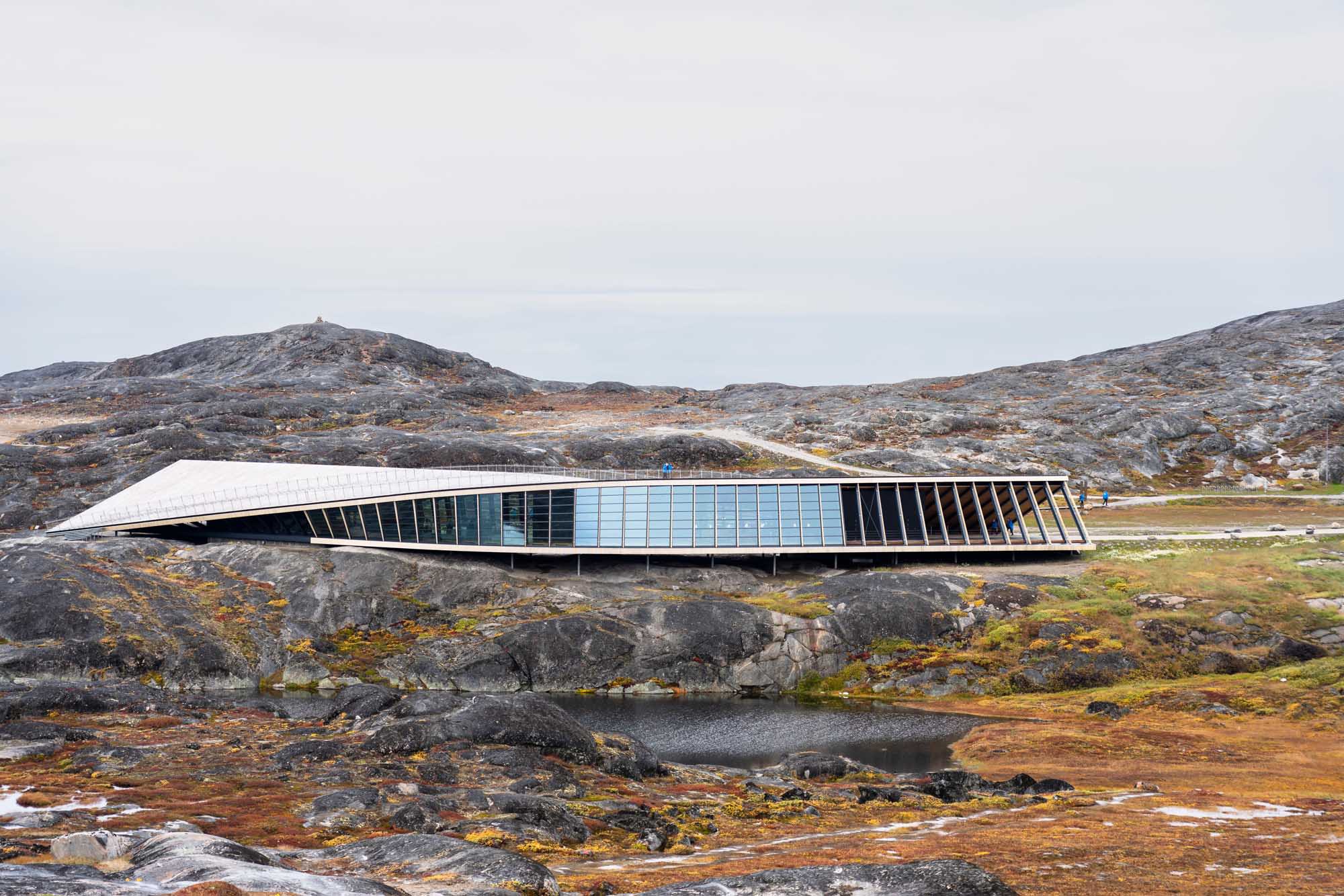 A striking contemporary building with a curved white roof and extensive blue-tinted glass panels sits atop rocky Arctic terrain in Greenland. The structure's sleek design contrasts dramatically with the barren, moss-covered landscape and distant mountains, exemplifying innovative architecture in one of the world's most remote regions.