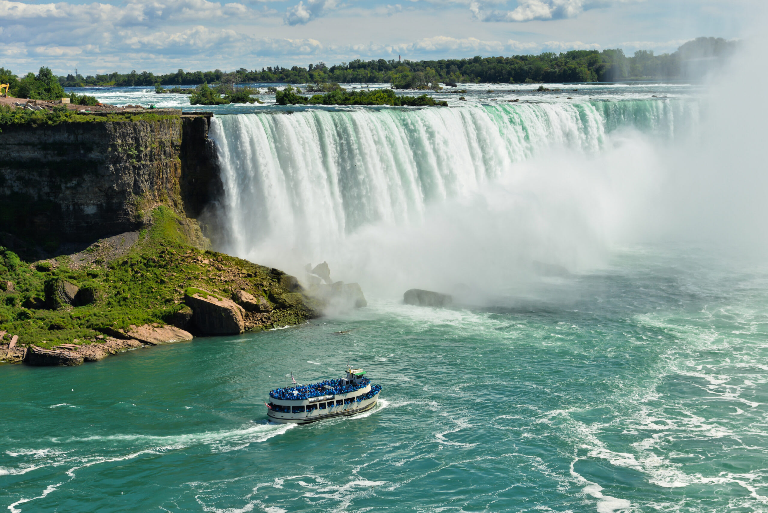 A scenic view of Niagara Falls' Horseshoe Falls with a passenger tour boat navigating the turquoise waters below, surrounded by mist and lush greenery on the Canadian side.
