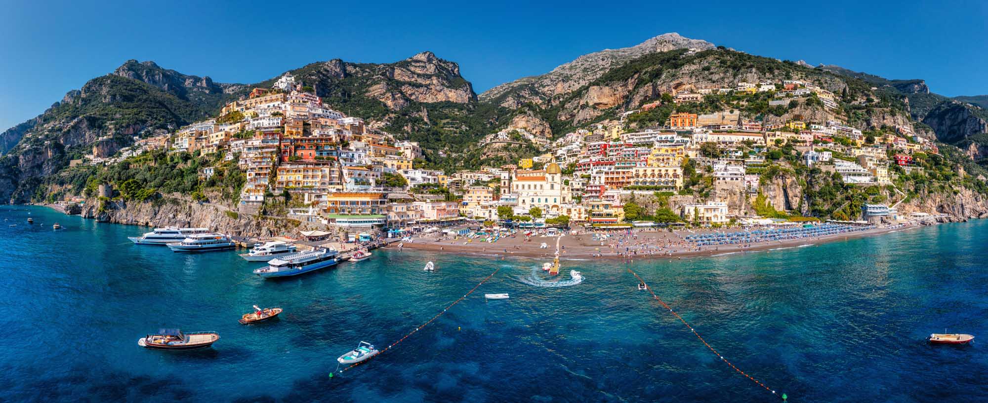 An aerial panoramic view of Positano, a picturesque cliffside village perched dramatically on the Amalfi Coast in Italy. The colorful cascading buildings descend steeply toward a sandy beach and turquoise Mediterranean waters dotted with boats, showcasing the iconic architecture and stunning coastal landscape characteristic of Campania.