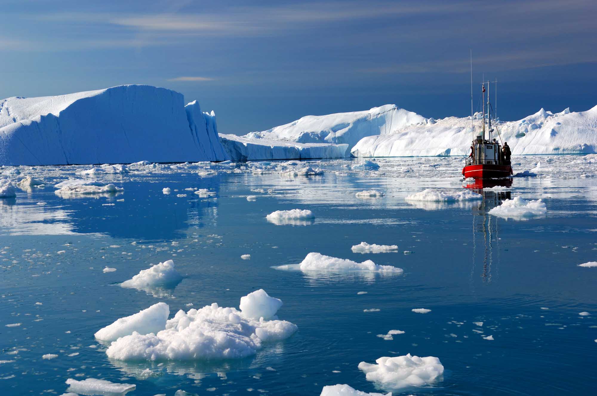 A red and black research vessel navigates through icy waters surrounded by massive tabular icebergs