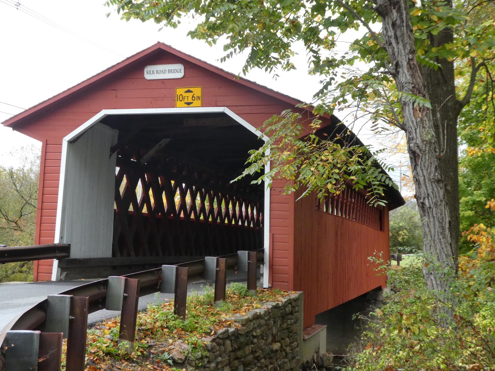 Silk Road Covered Bridge in Autumn The historic Silk Road covered bridge, a red wooden structure with white trim and distinctive latticed interior, spanning across a stone foundation. The bridge features a clearance height of 10 feet 6 inches and is surrounded by autumn foliage and mature trees.