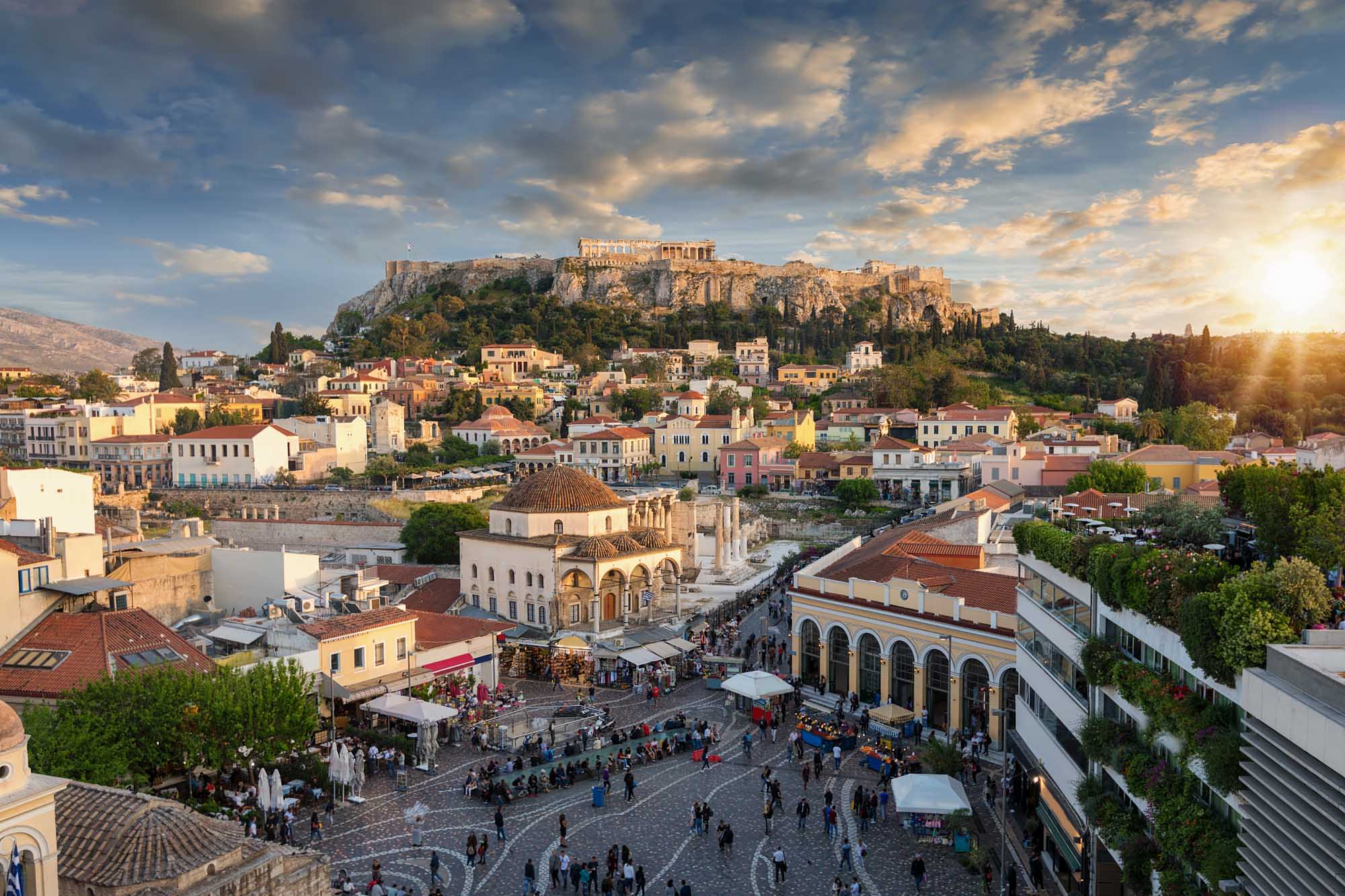 A stunning golden-hour view of Athens' historic Plaka district with colorful neoclassical buildings clustered below the illuminated Acropolis and Parthenon Temple perched atop a rocky hill.
