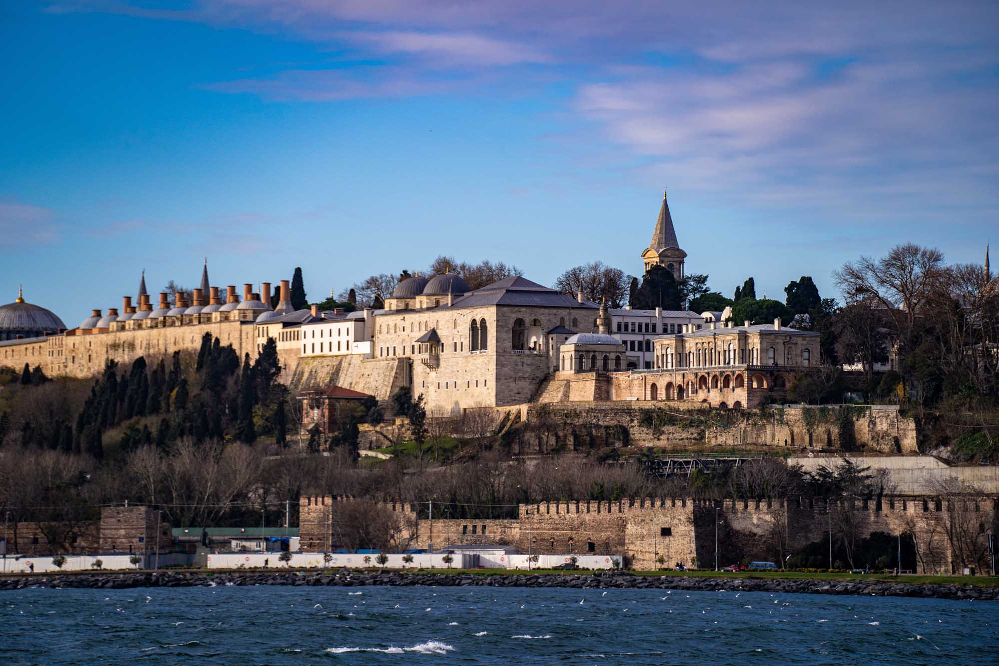Topkapi Palace View from the Bosphorus A stunning waterfront view of the historic Topkapi Palace in Istanbul, Turkey, photographed from the Bosphorus Strait. The ornate Ottoman architecture features distinctive domes, minarets, and stone fortifications set against a clear blue sky, with cypress trees framing the palatial complex on the peninsula.