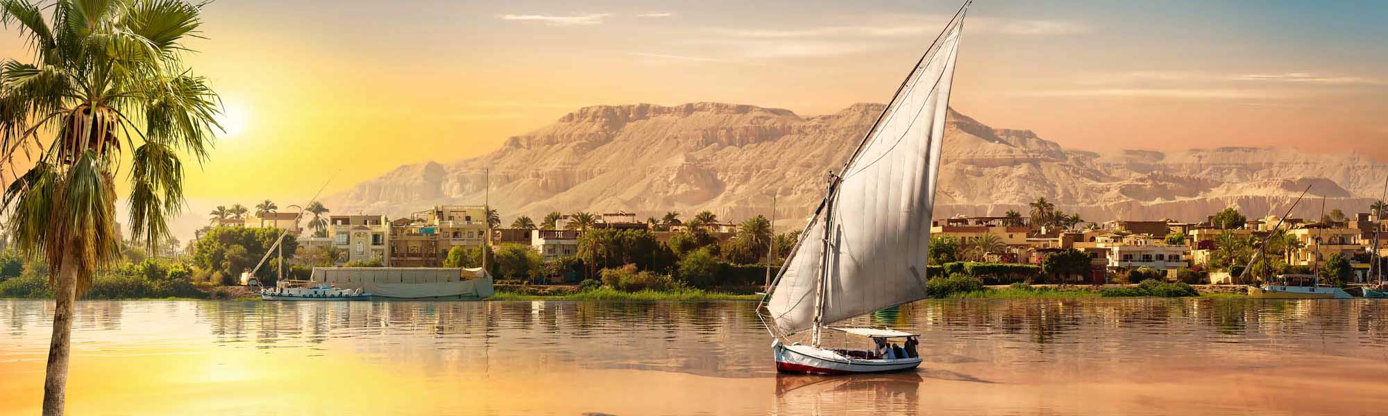 A traditional Egyptian felucca sailboat with white canvas sails glides peacefully across the Nile River at golden sunrise, with palm-lined riverbanks, colorful buildings, and desert cliffs in the background.
