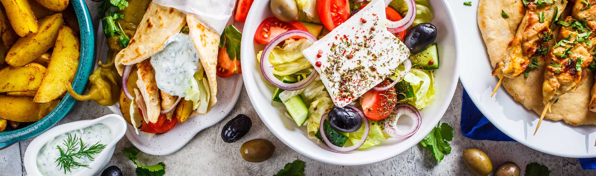 A vibrant overhead display of authentic Greek dishes including Greek salad with feta cheese and olives, grilled chicken souvlaki skewers, gyros wraps with tzatziki sauce, and crispy baked potato wedges, arranged on a gray surface with fresh herbs and garnishes.