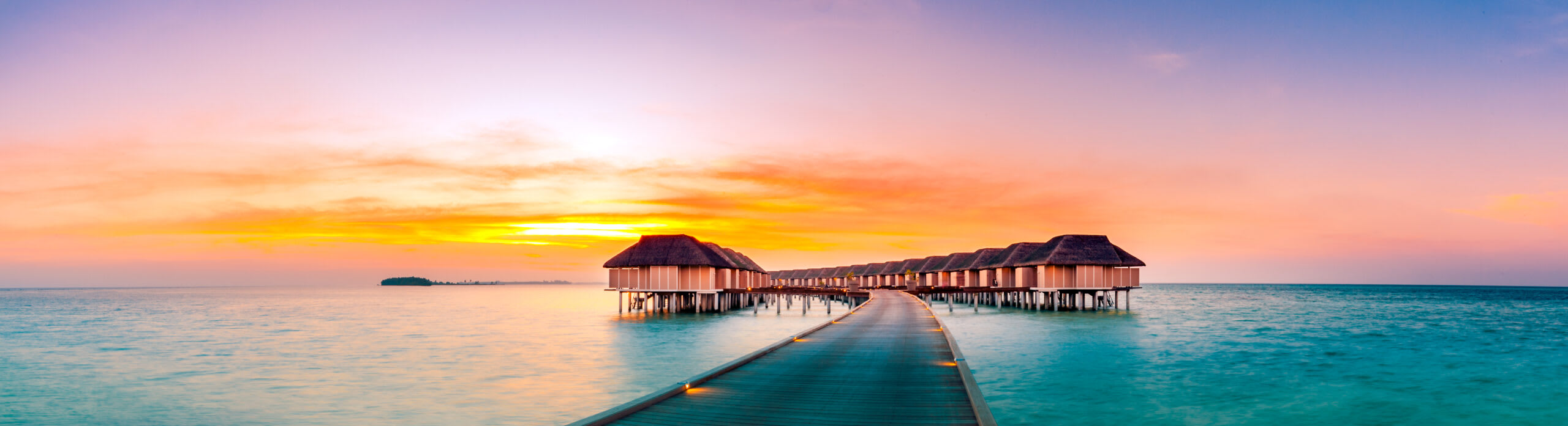 Tropical Overwater Bungalows at Sunset A serene tropical resort featuring traditional thatched-roof overwater bungalows connected by wooden walkways, set in crystal-clear turquoise waters during a spectacular golden sunset with warm orange and pink sky reflections.