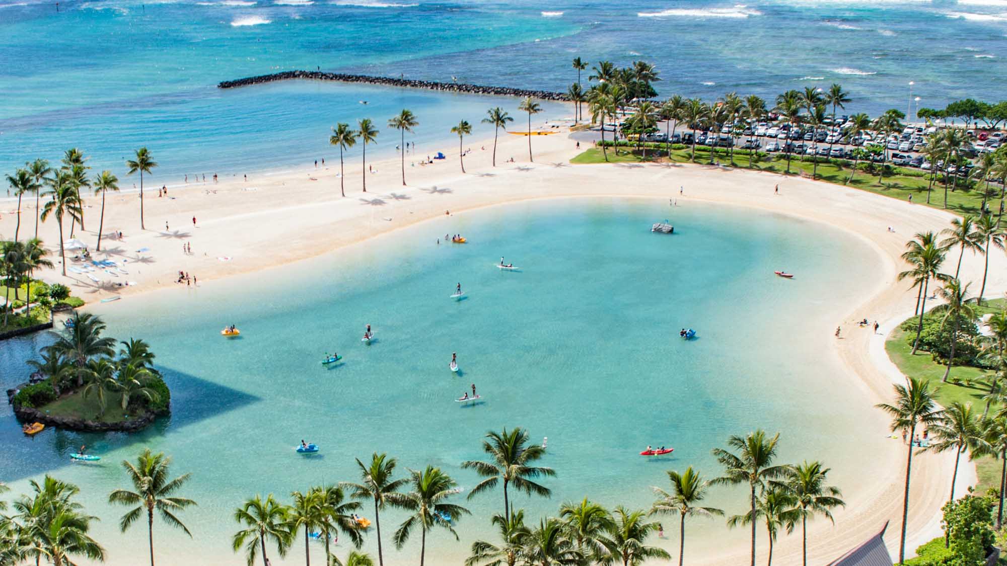 An aerial view of a pristine tropical beach destination featuring a stunning turquoise lagoon with soft white sand, scattered palm trees, and numerous water activities.