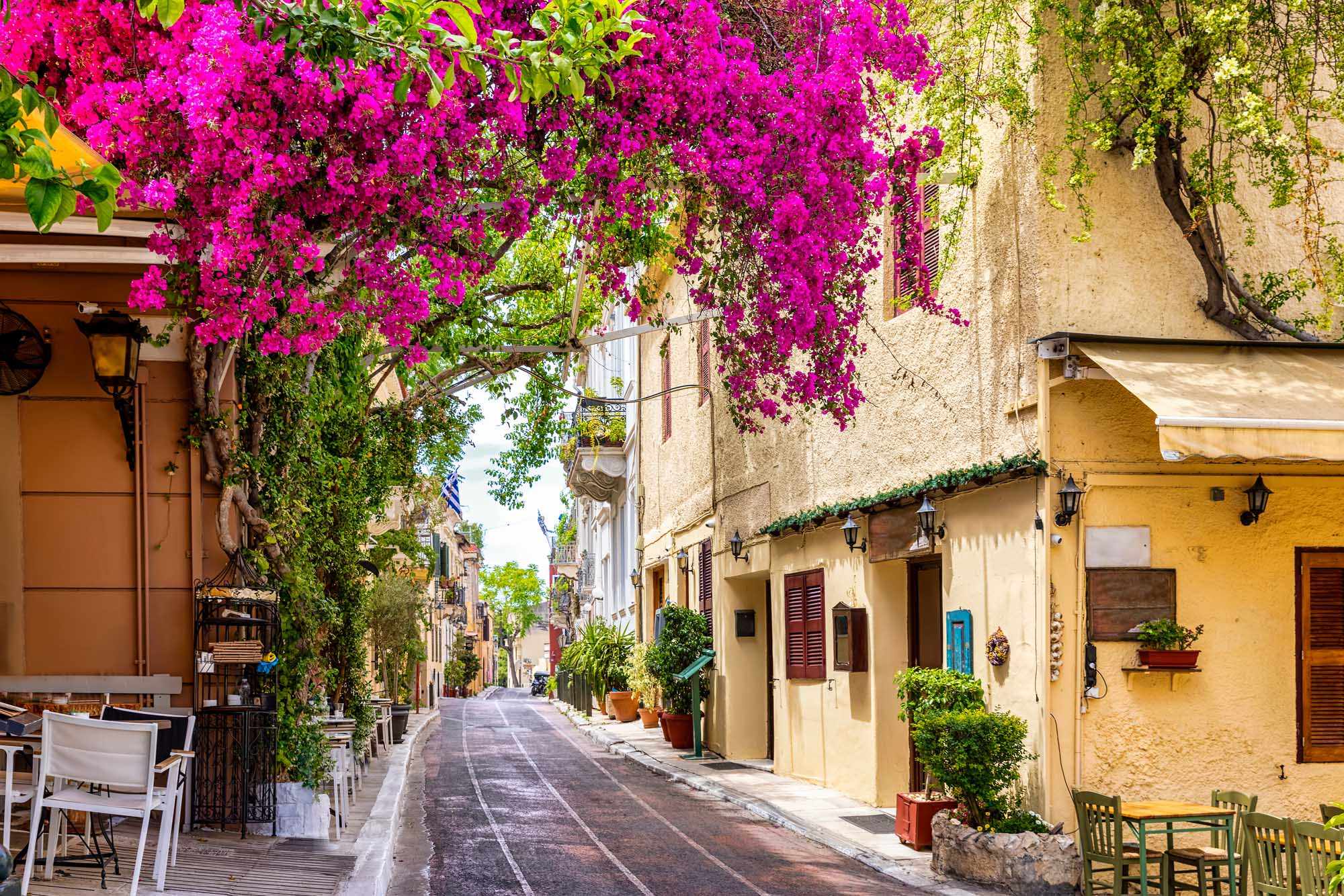 A charming Mediterranean alleyway lined with traditional stone buildings and cascading magenta bougainvillea flowers overhead, featuring characteristic architecture with colorful doors, shutters, and outdoor cafe seating along a quiet street.