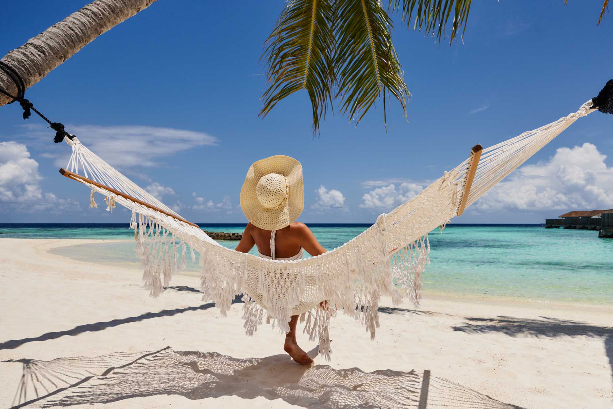 A rear view of a woman wearing a straw sun hat relaxing in a white macrame hammock strung between palm trees on a pristine tropical beach, overlooking turquoise waters and a clear blue sky dotted with white clouds.