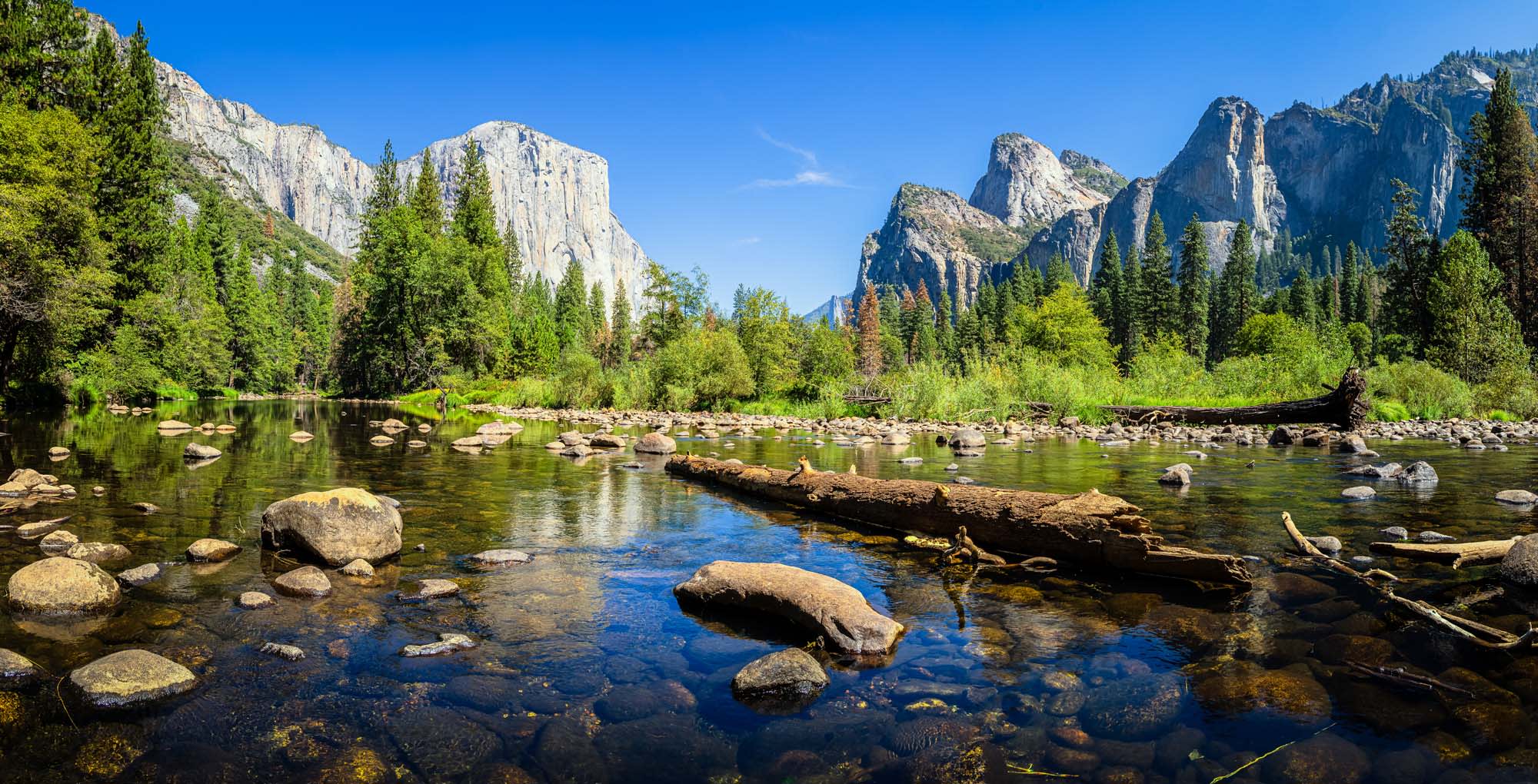 Yosemite Valley with El Capitan and Merced River A stunning panoramic view of Yosemite Valley featuring the iconic granite cliffs of El Capitan and Three Brothers formations rising majestically above a pristine stretch of the Merced River. The scene captures clear reflections in the calm water with scattered boulders, dense coniferous forest, and a brilliant blue summer sky.