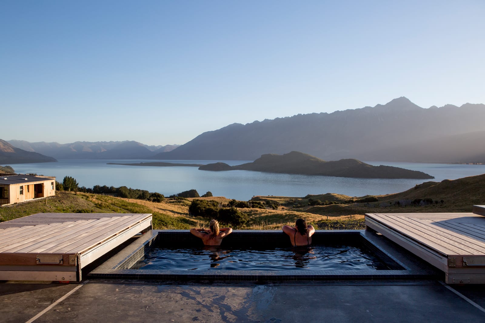 Two people relaxing in infinity pool overlooking mountain lake and forested landscape at sunset