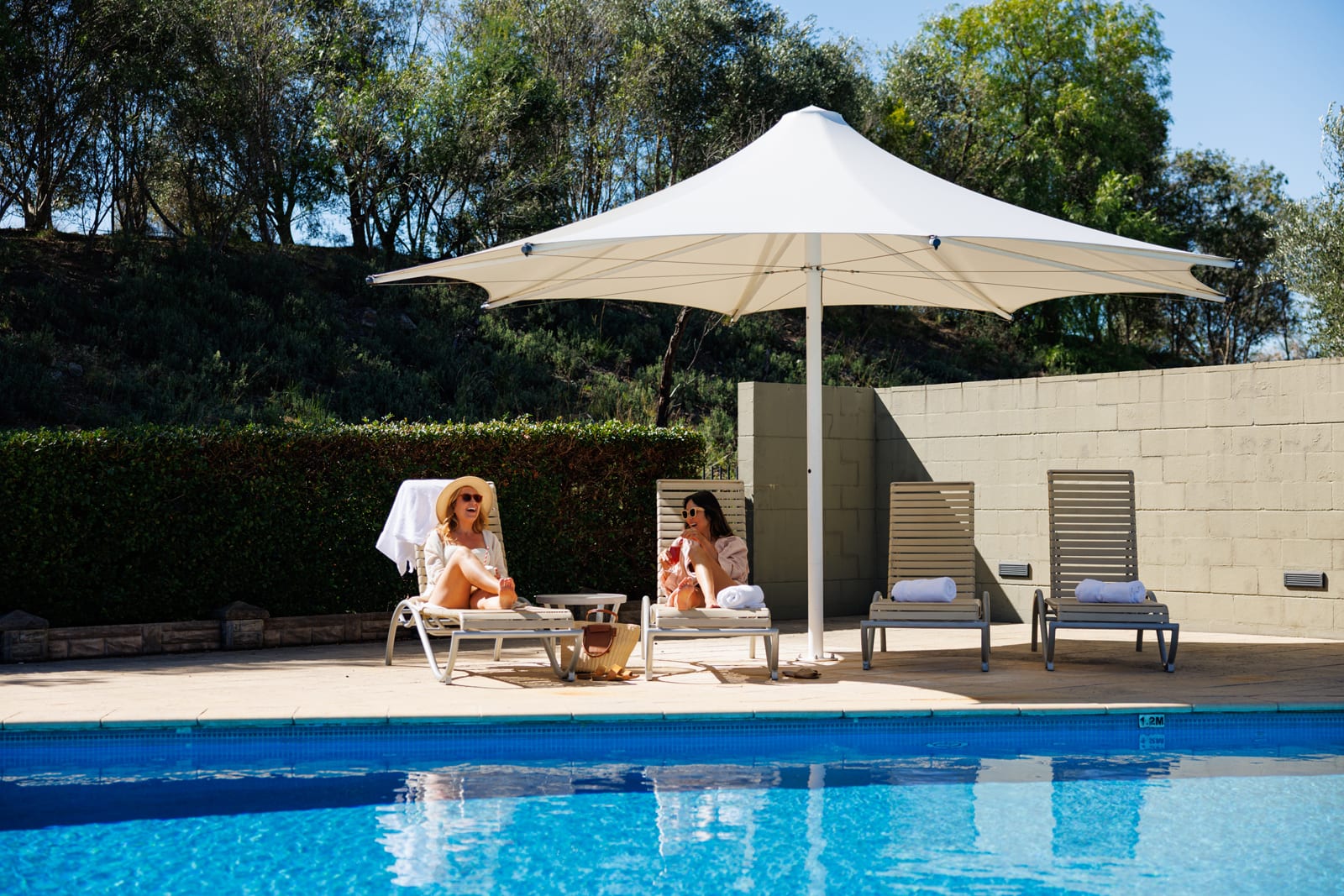 Elysia Wellness Retreat_Pool2 Two women relaxing on lounge chairs by a swimming pool under a white canopy umbrella