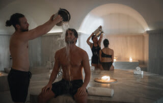 Man pouring water over person's head in traditional hammam bath with others in background