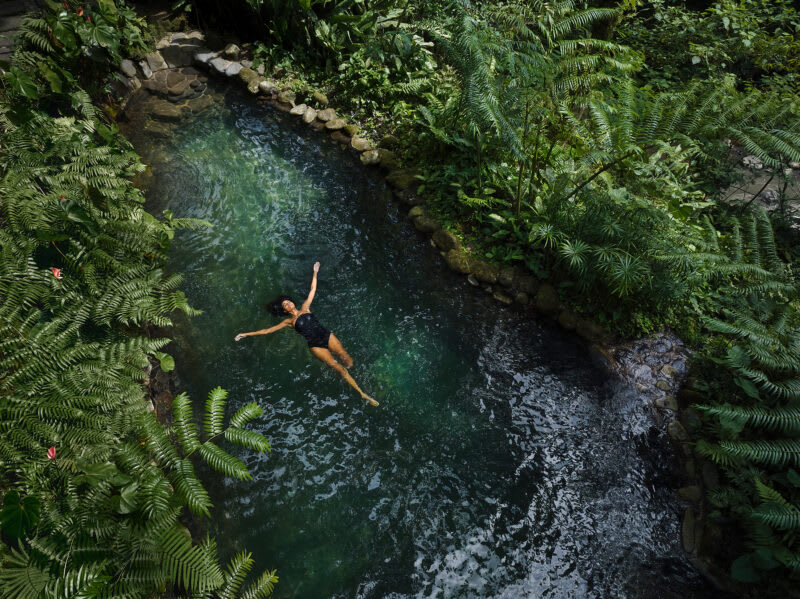 COMO-Shambhala-Estate-3-1 A person floating peacefully in a lush green jungle stream surrounded by ferns and tropical vegetation. The water is a deep emerald green with stone-lined banks.