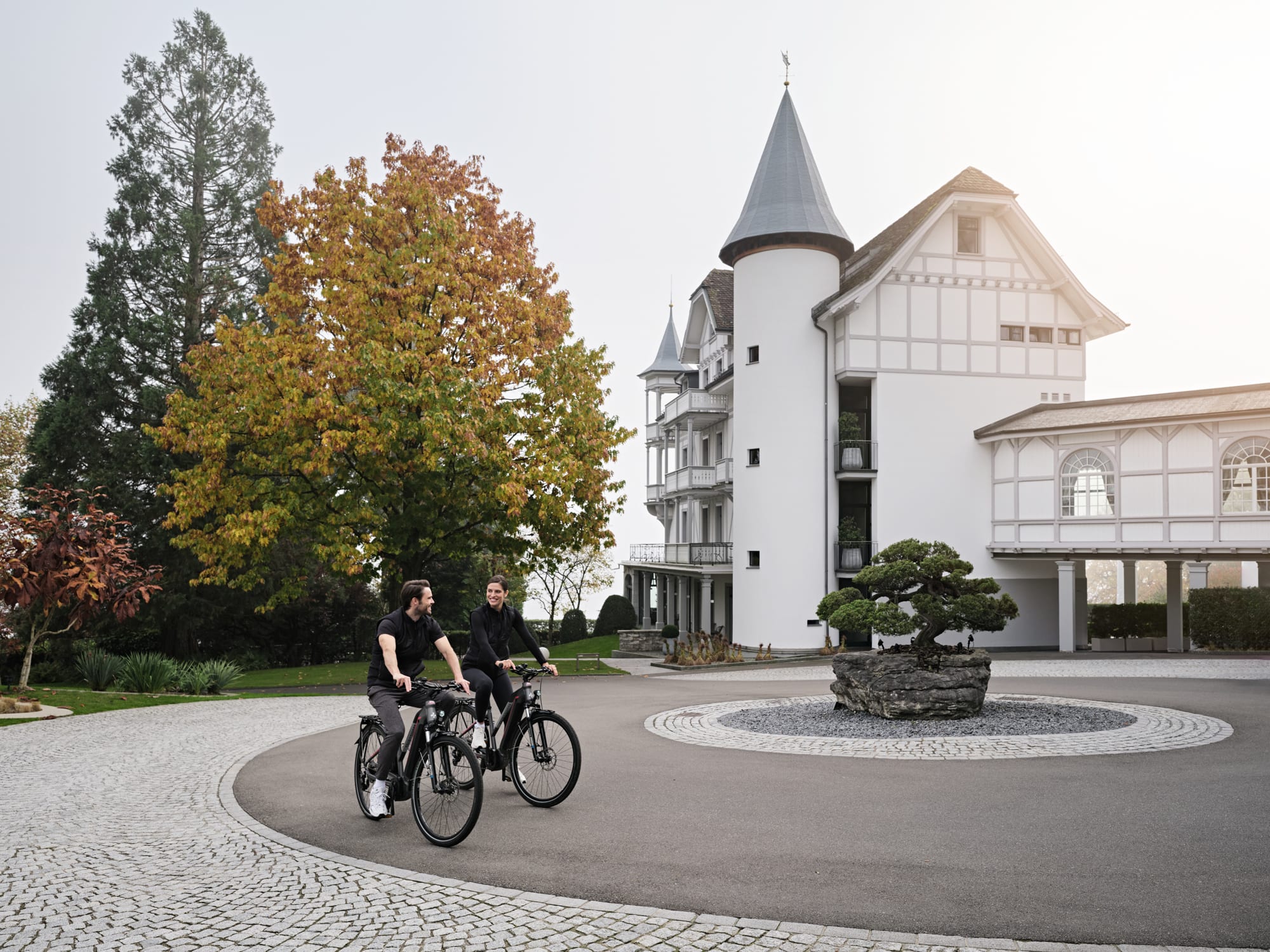 GGArchard_Chenot_2024-10_06 Two cyclists riding bikes on circular driveway in front of white castle-like mansion with turrets and autumn trees