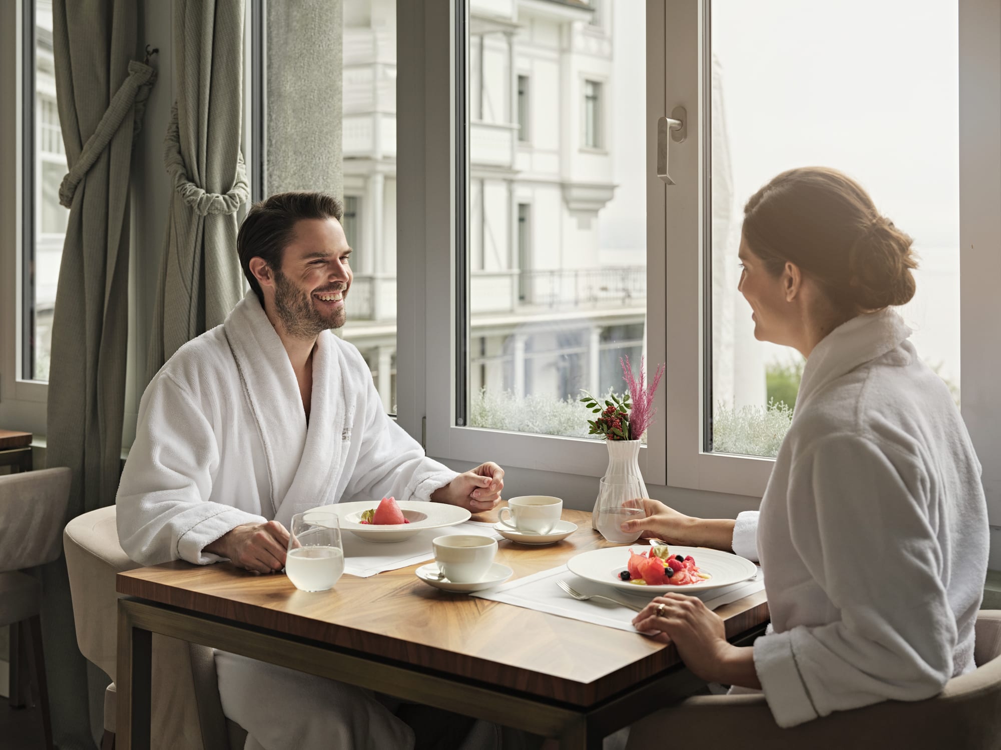 GGArchard_Chenot_2024-10_10 Couple in spa robes enjoying breakfast at a table by large windows overlooking a city
