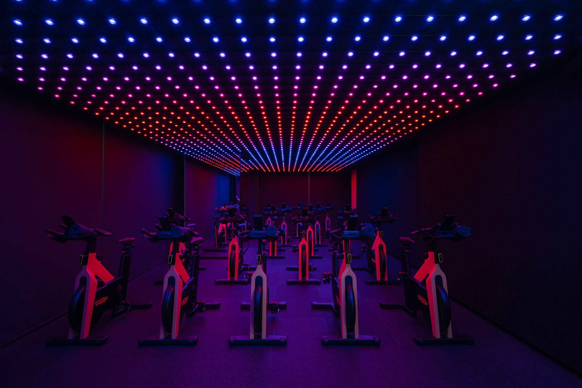 Siro One Zabeel, Siro Hotels Modern spin studio with red stationary bikes arranged in rows beneath vibrant LED ceiling lights in blue, purple, and orange gradient.