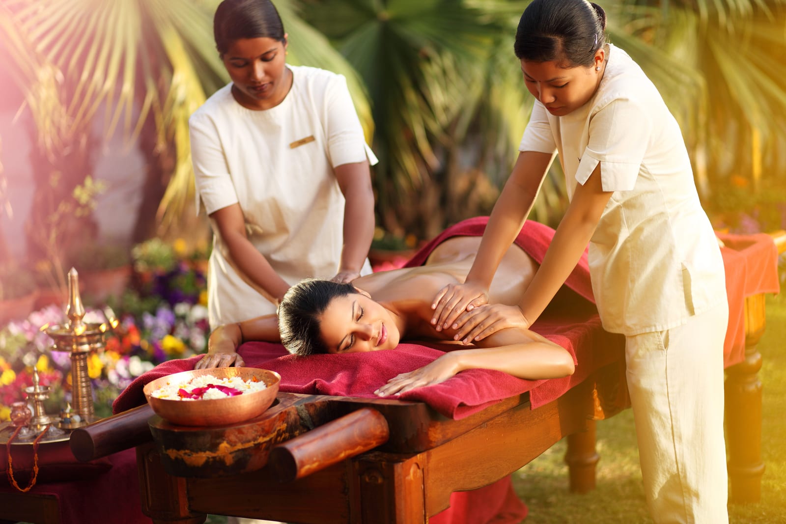 Wellness_Ayurveda_therapy 1 Two spa therapists performing back massage on woman in tropical garden setting with flowers and oil bowl