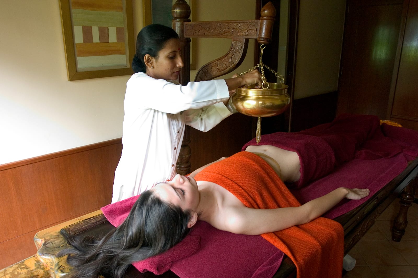 Wellness_Ayurveda_therapy 5 Ayurvedic practitioner performing oil treatment on patient lying on massage table with brass vessel