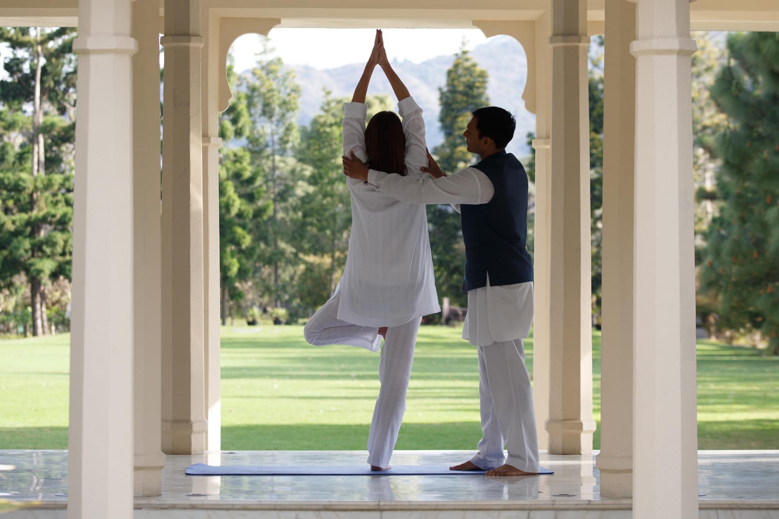 Wellness_Yoga 3 Yoga instructor guiding student in tree pose under columned pavilion with mountain view