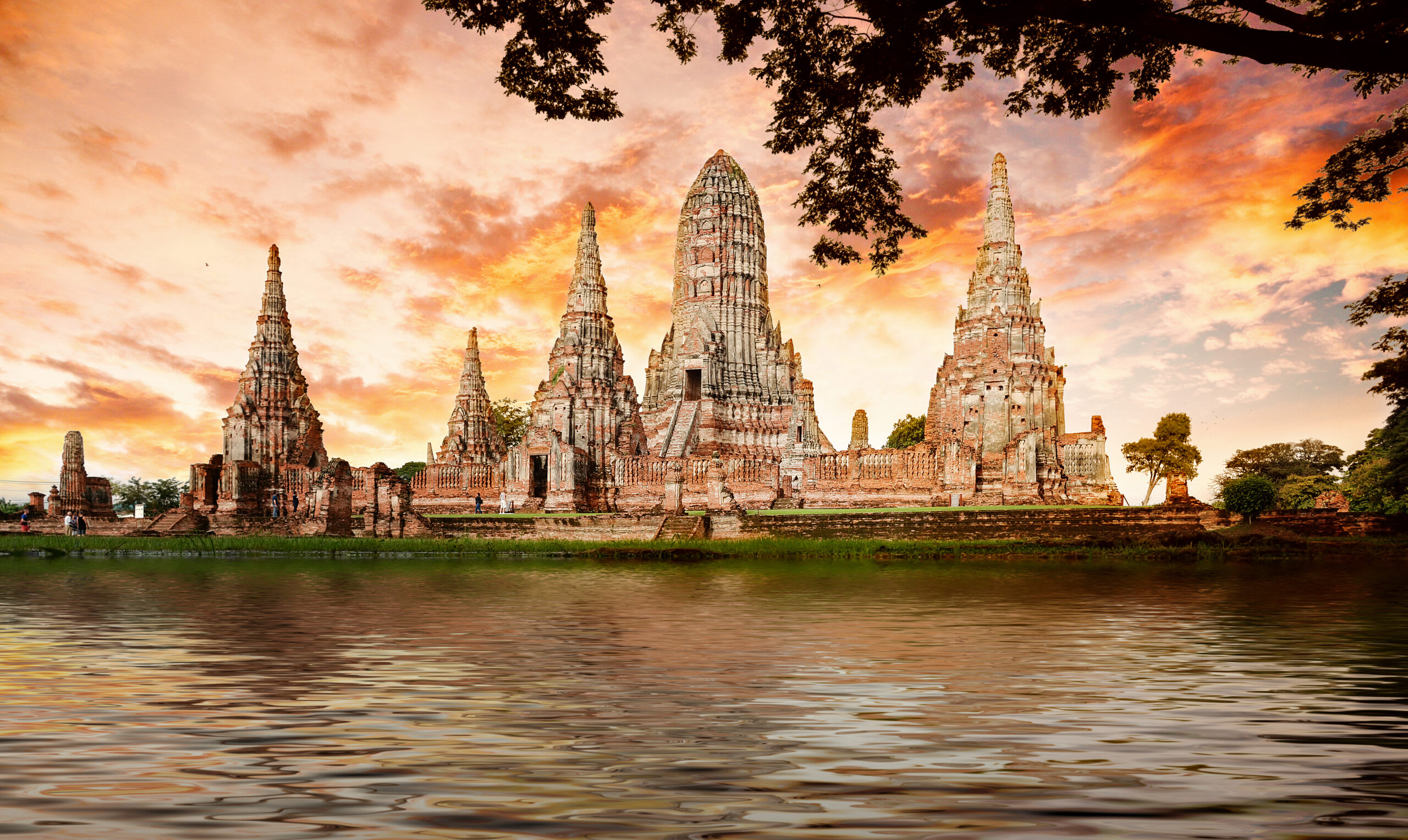 Ancient Thai temple ruins with ornate spires reflected in river at sunset