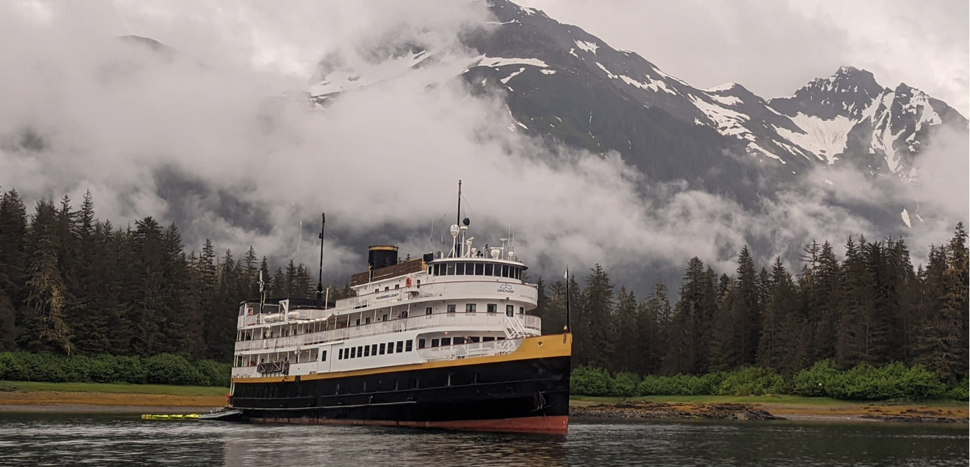 A white and yellow passenger ferry navigates through calm waters near a forested shoreline