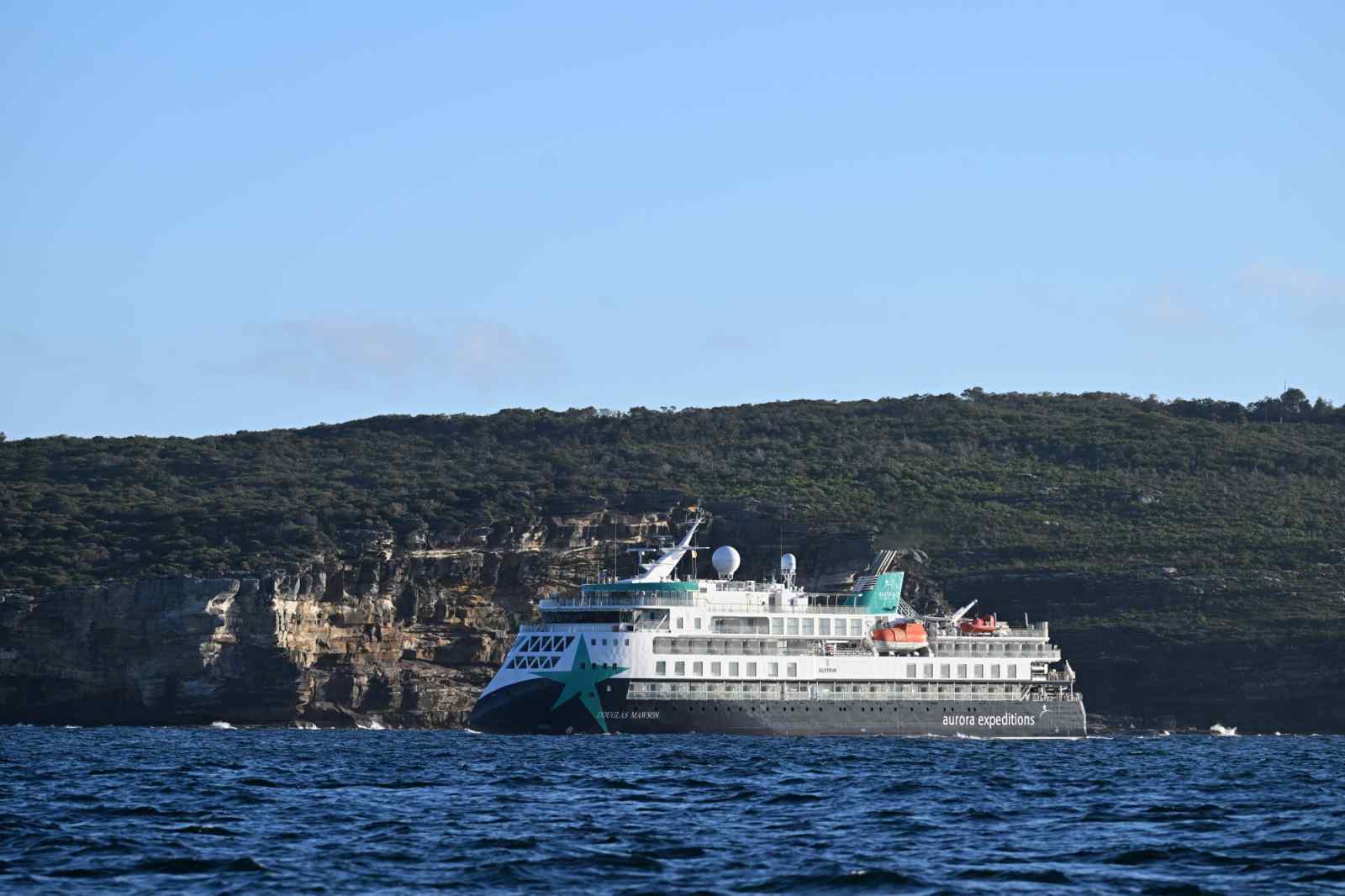 An Aurora Expeditions expedition cruise ship sailing close to a rugged, rocky coastline with dense