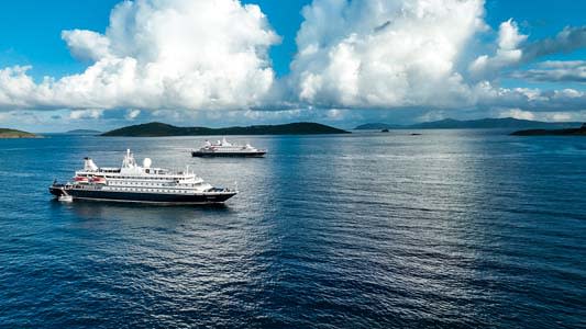 Two white cruise ships anchored in a serene blue ocean with dramatic cloudy sky and mountainous