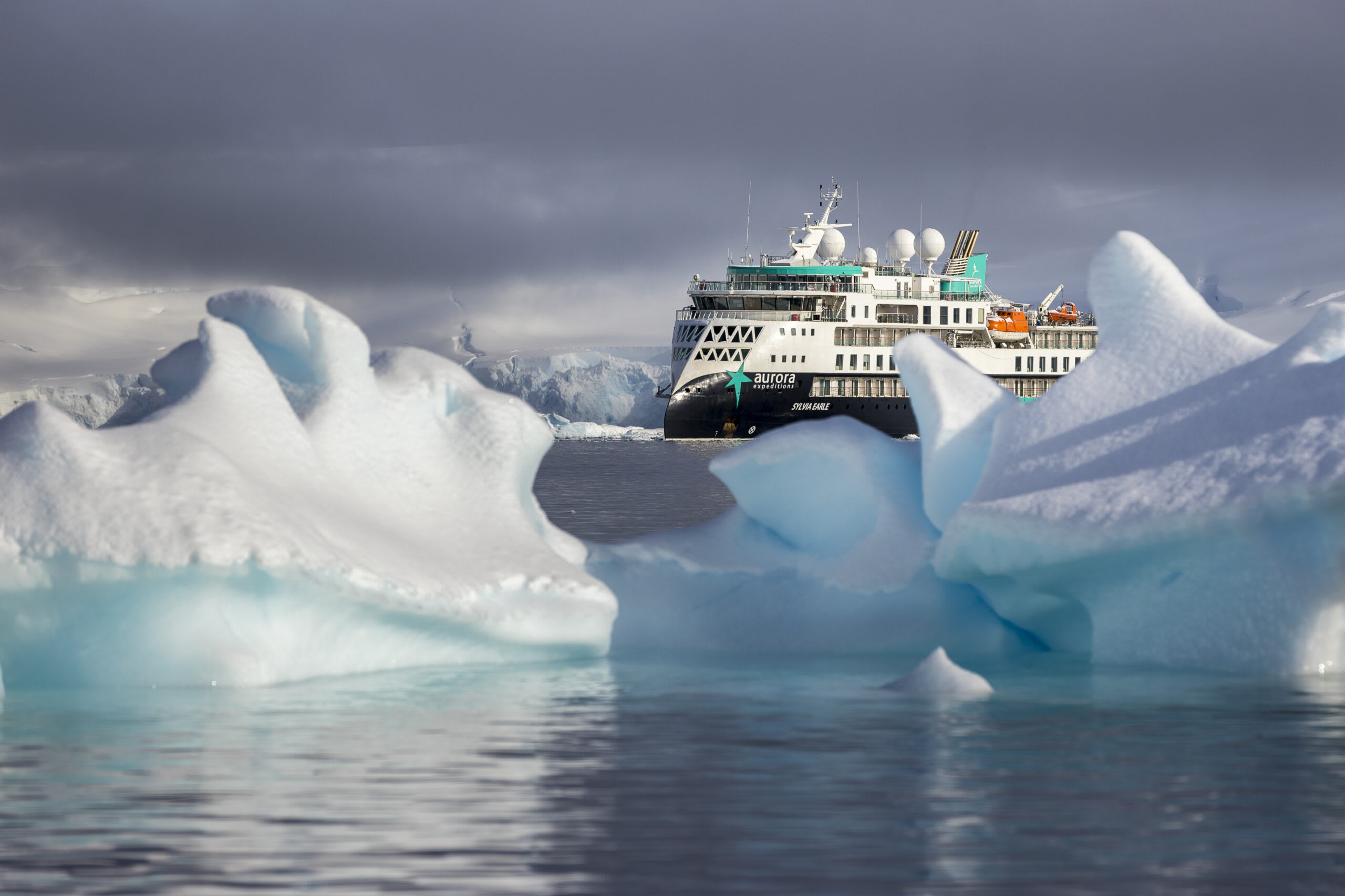 A white and teal expedition cruise ship navigating through blue-white icebergs in a misty polar seascape with dramatic cloudy skies.