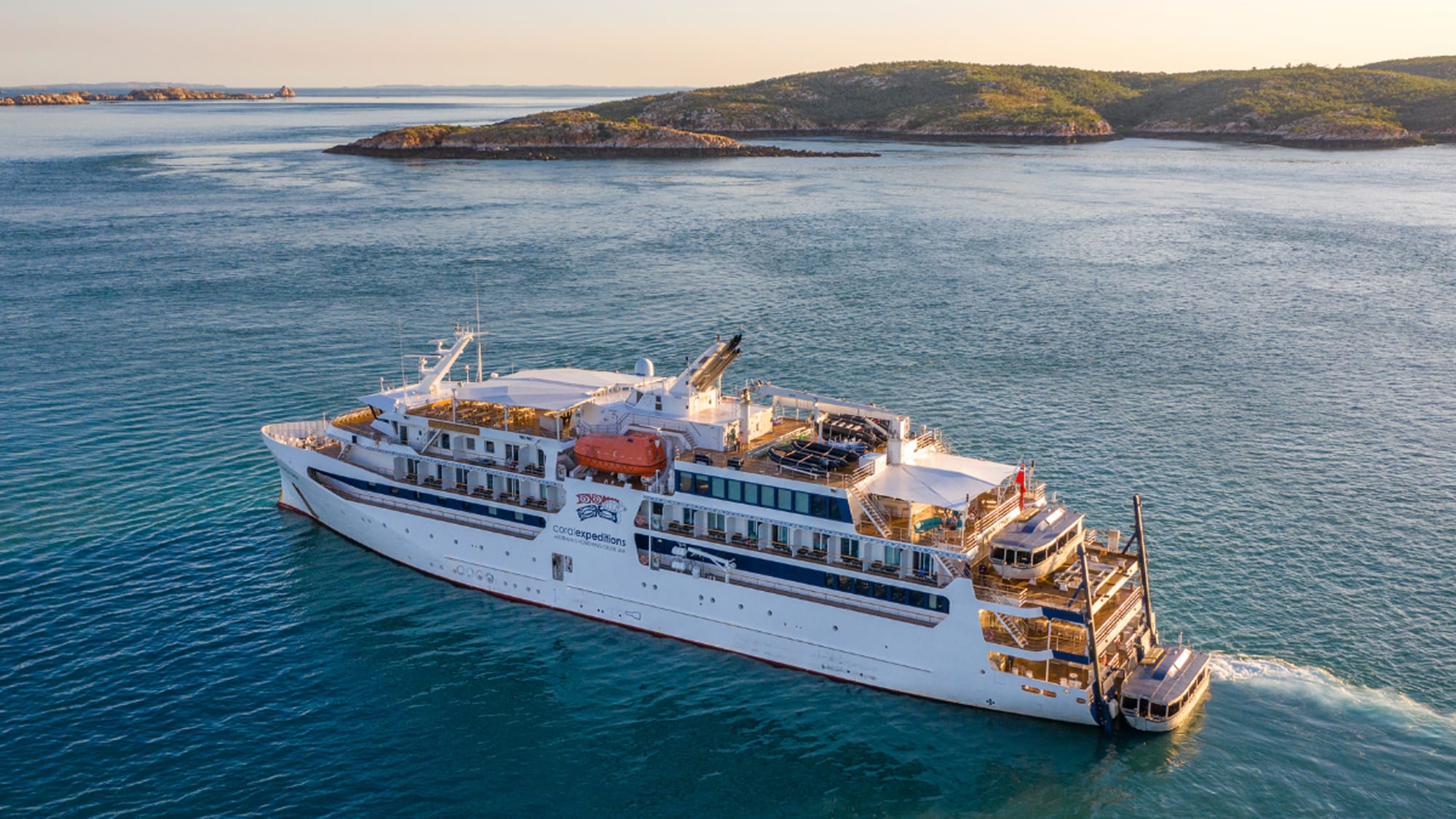 A white expedition-style cruise ship sailing through blue waters near a rocky, forested coastline