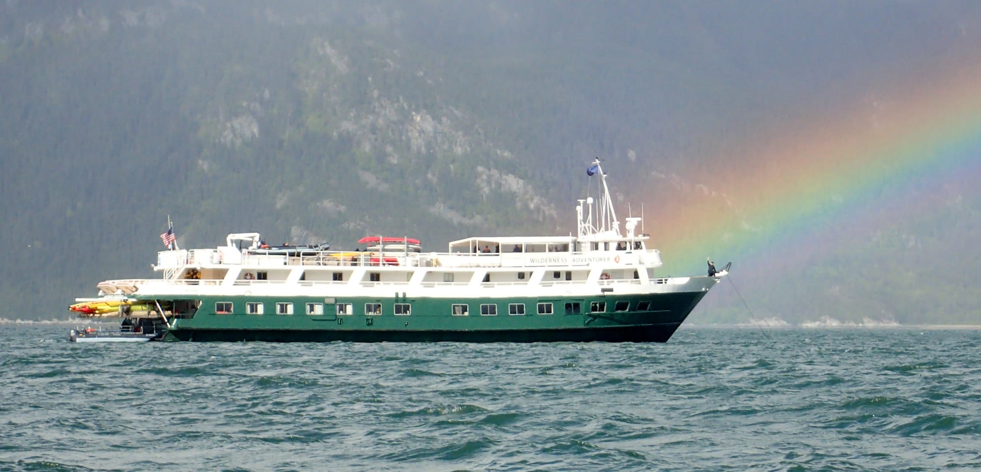 A green and white expedition or cruise ship sailing on choppy waters with a vivid rainbow arching
