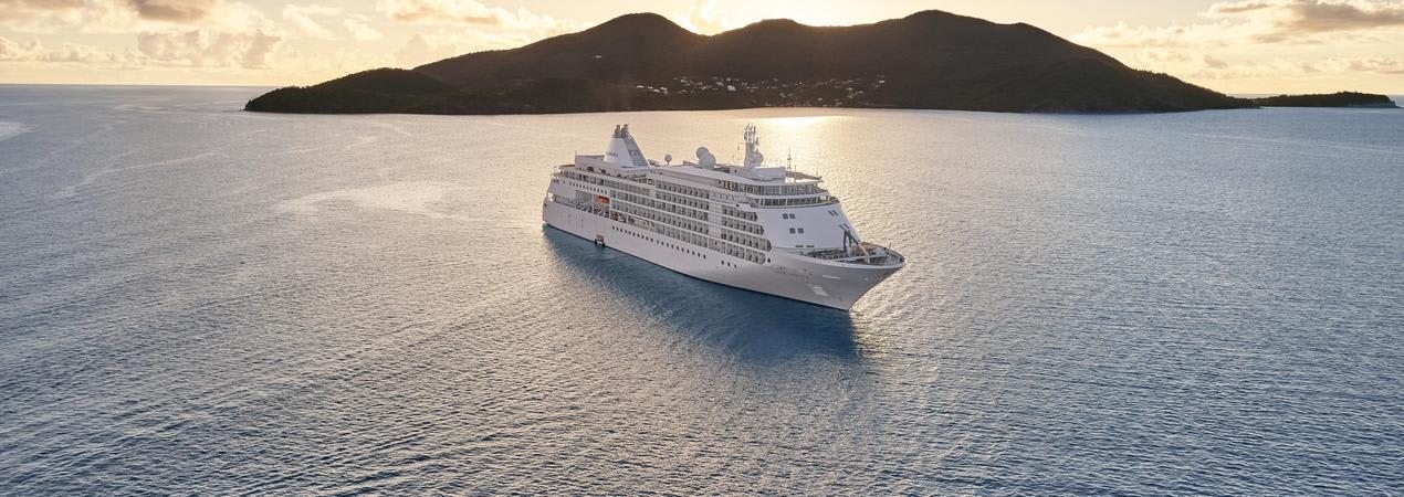 A white luxury cruise ship anchored near a mountainous island during golden hour, with calm waters