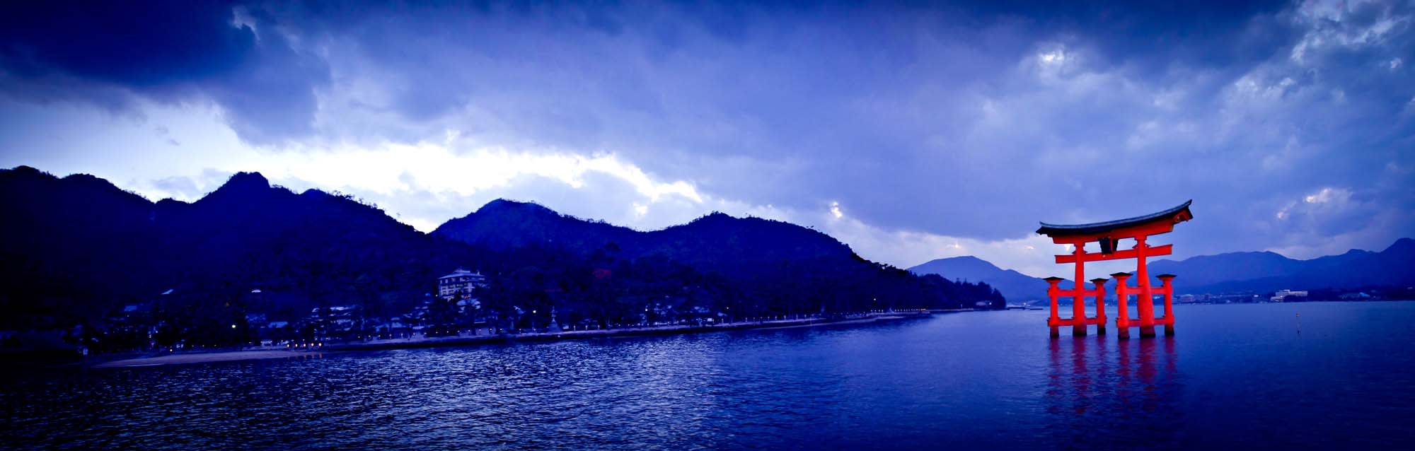 A vibrant red torii gate stands in the blue waters of Miyajima, with misty mountains and a coastline in the background during a serene twilight hour.