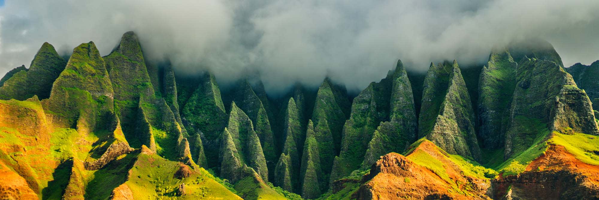 Dramatic mountainous coastline with sharp, green-covered peaks rising dramatically against a misty cloudy background, showcasing the rugged terrain of Kauai, Hawaii.