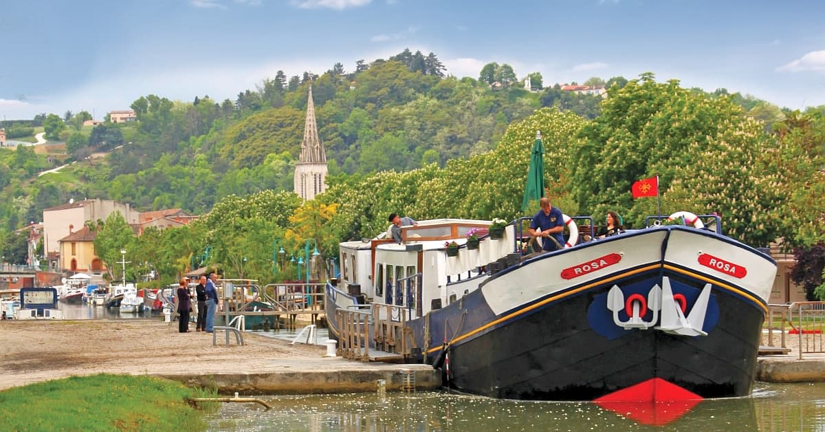 River Cruise Boat on European Waterway The river cruise ship Rosa is moored at a picturesque European riverport with passengers on deck, surrounded by lush green hillsides, a church spire visible in the background, and traditional riverside architecture. The scene captures the essence of leisurely river cruising through a charming continental landscape.