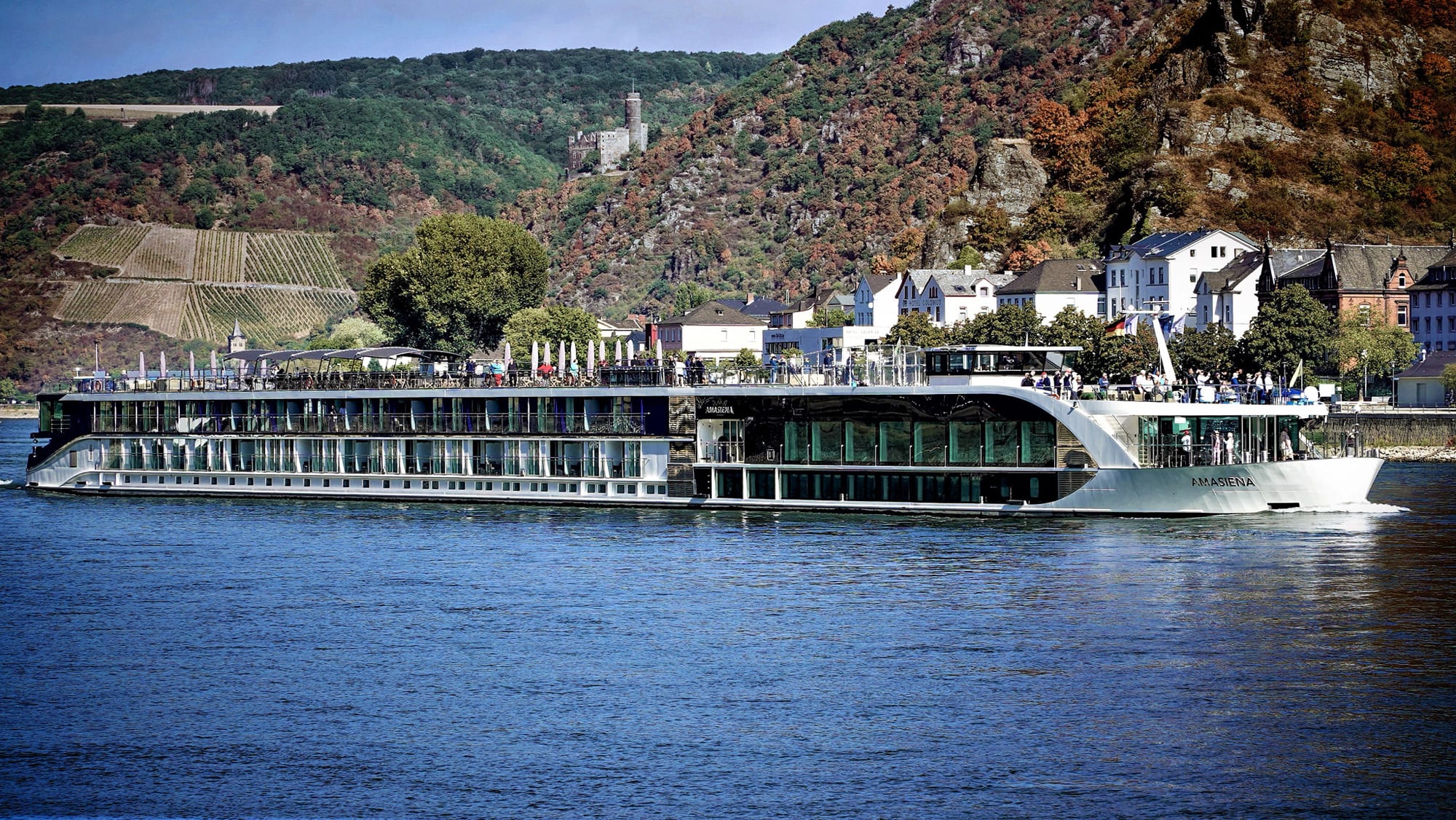 A modern river cruise ship named Amasiena docked along a picturesque riverside town with vineyards
