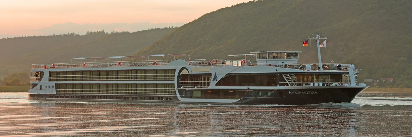 A modern river cruise vessel sailing on a calm river with forested hills in the background during