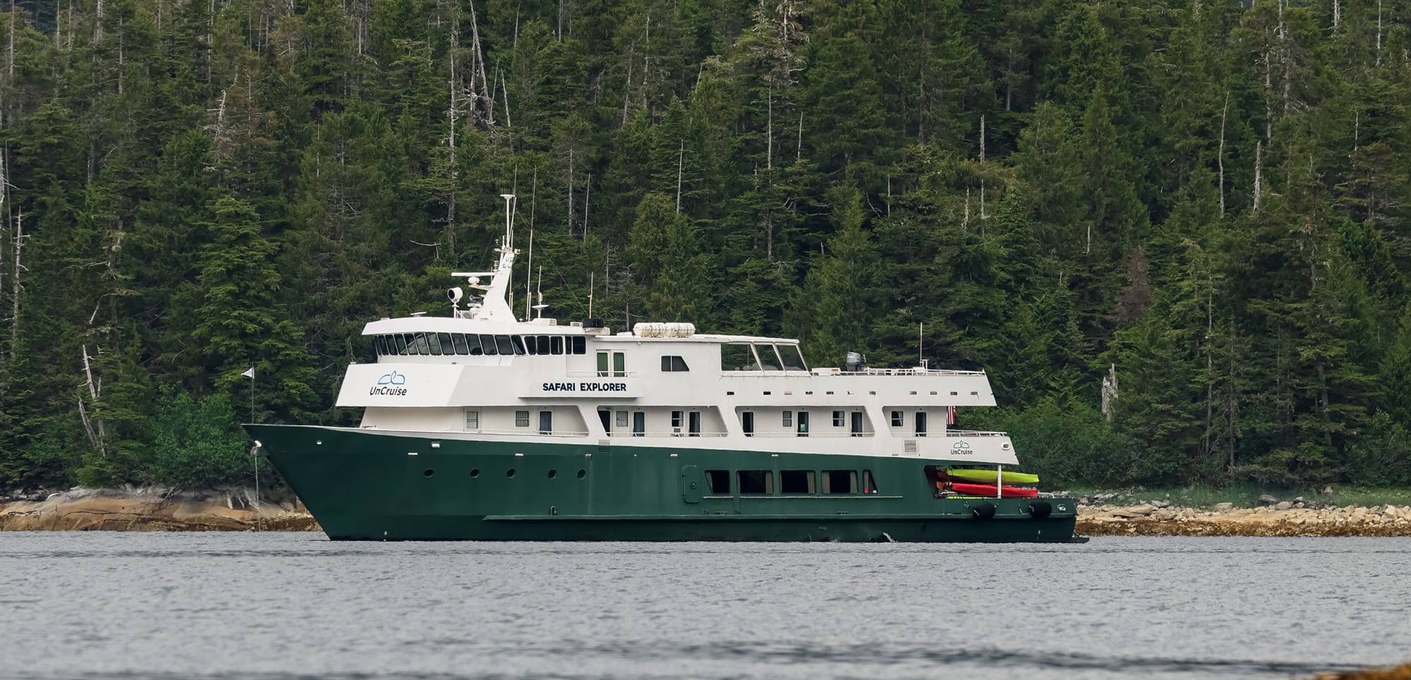 Safari Explorer expedition cruise ship anchored near evergreen forest coastline with colorful kayaks