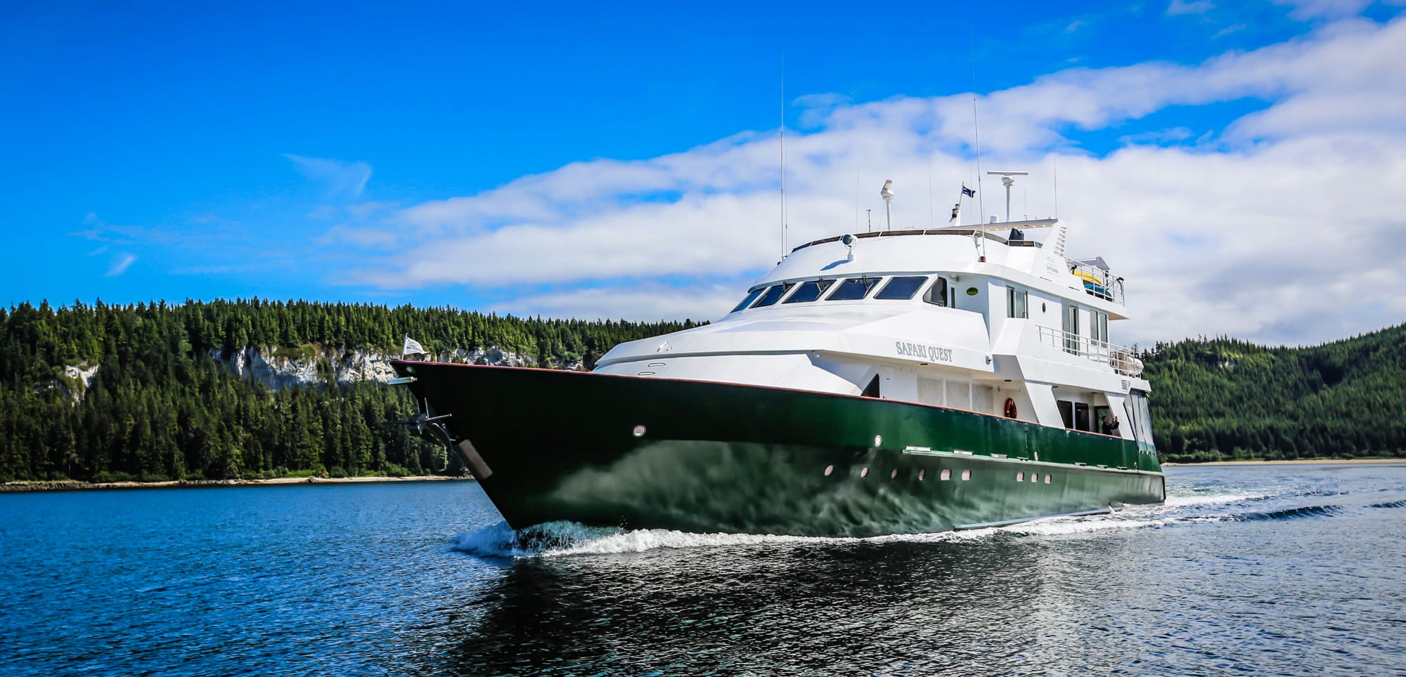 A white and green expedition cruise ship named Safari Quest navigating through a scenic forested
