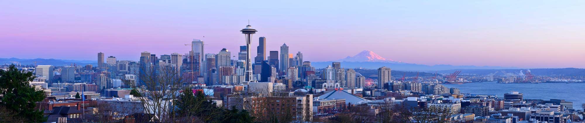 A panoramic view of Seattle's downtown skyline featuring the iconic Space Needle, with Mount Rainier in the background during a pastel-colored twilight hour. The cityscape showcases modern skyscrapers, urban buildings, and the waterfront.