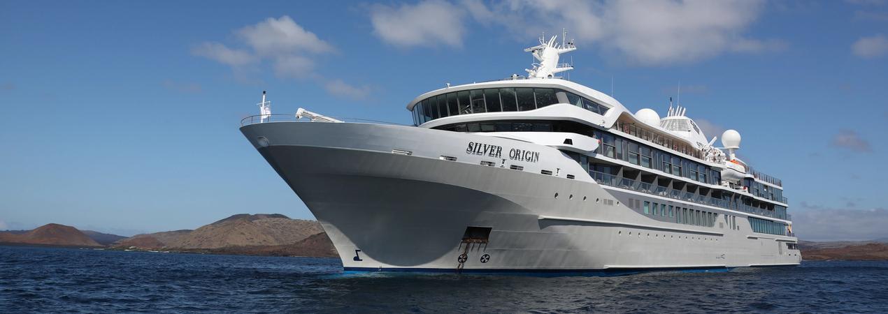 A sleek, white luxury cruise ship named Silver Origin sailing on blue waters with rocky islands