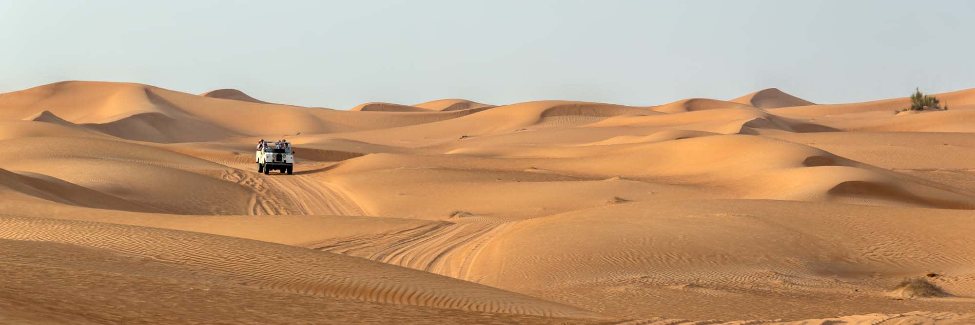 A vintage open-top 4x4 SUV traverses the golden sand dunes of the Dubai desert, navigating the undulating landscape with multiple sand dunes visible under clear skies. This captures the adventurous desert safari experience popular among tourists visiting the United Arab Emirates.