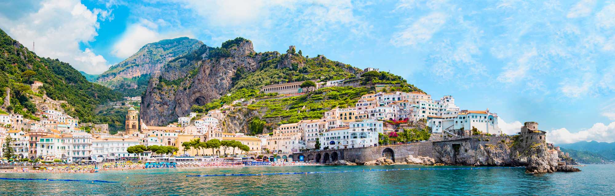 A stunning panoramic view of the picturesque Amalfi village nestled along dramatic cliffsides with colorful Mediterranean architecture, featuring pastel-colored houses cascading down the rocky coastline overlooking turquoise waters and a small sandy beach.