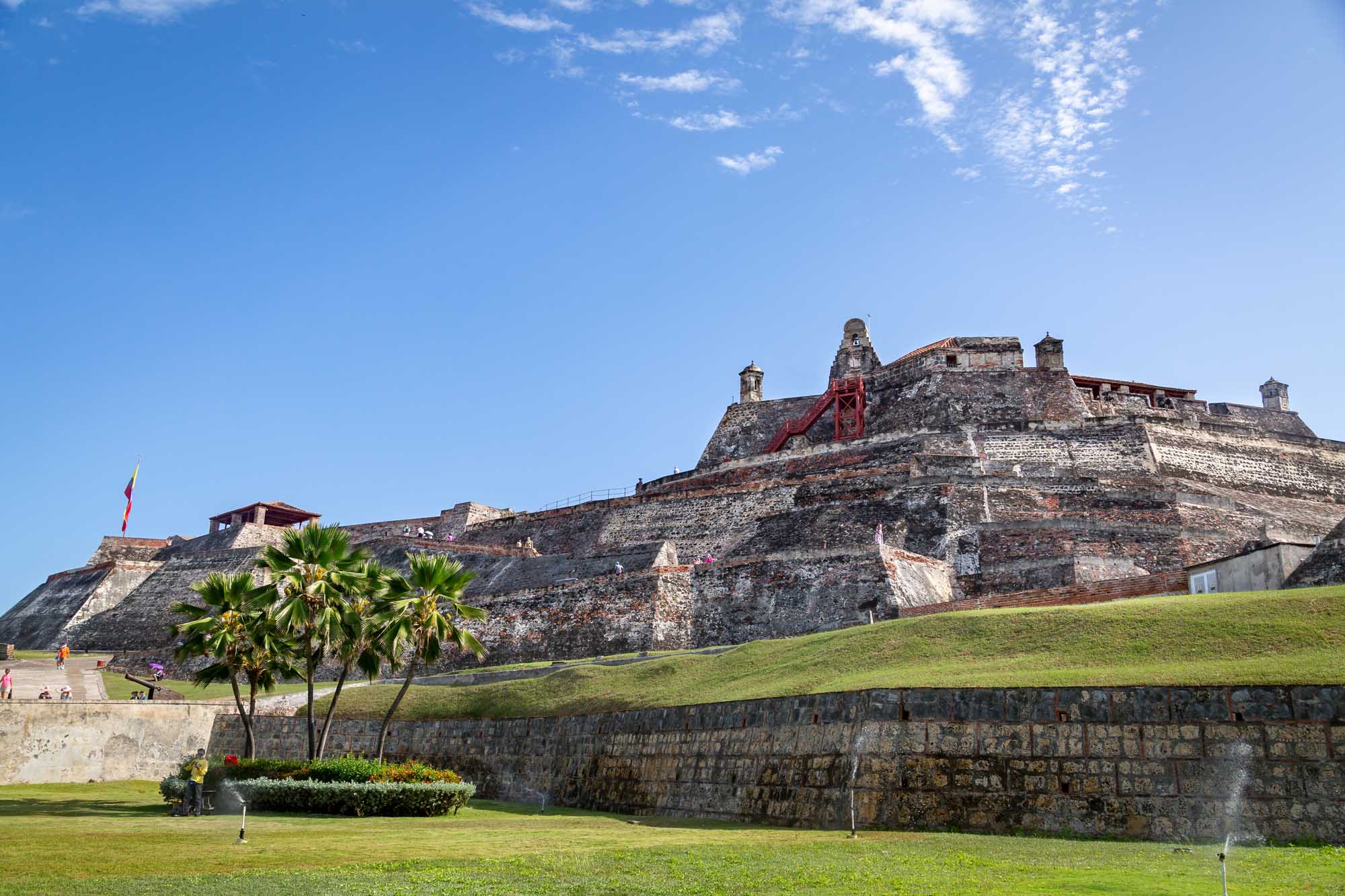 A UNESCO World Heritage Site fortress featuring distinctive red brick and stone ramparts with palm trees in the foreground, located in Cartagena, Colombia. The imposing 16th-17th century military structure showcases Spanish colonial architecture with its characteristic angled bastions and weathered fortification walls.
