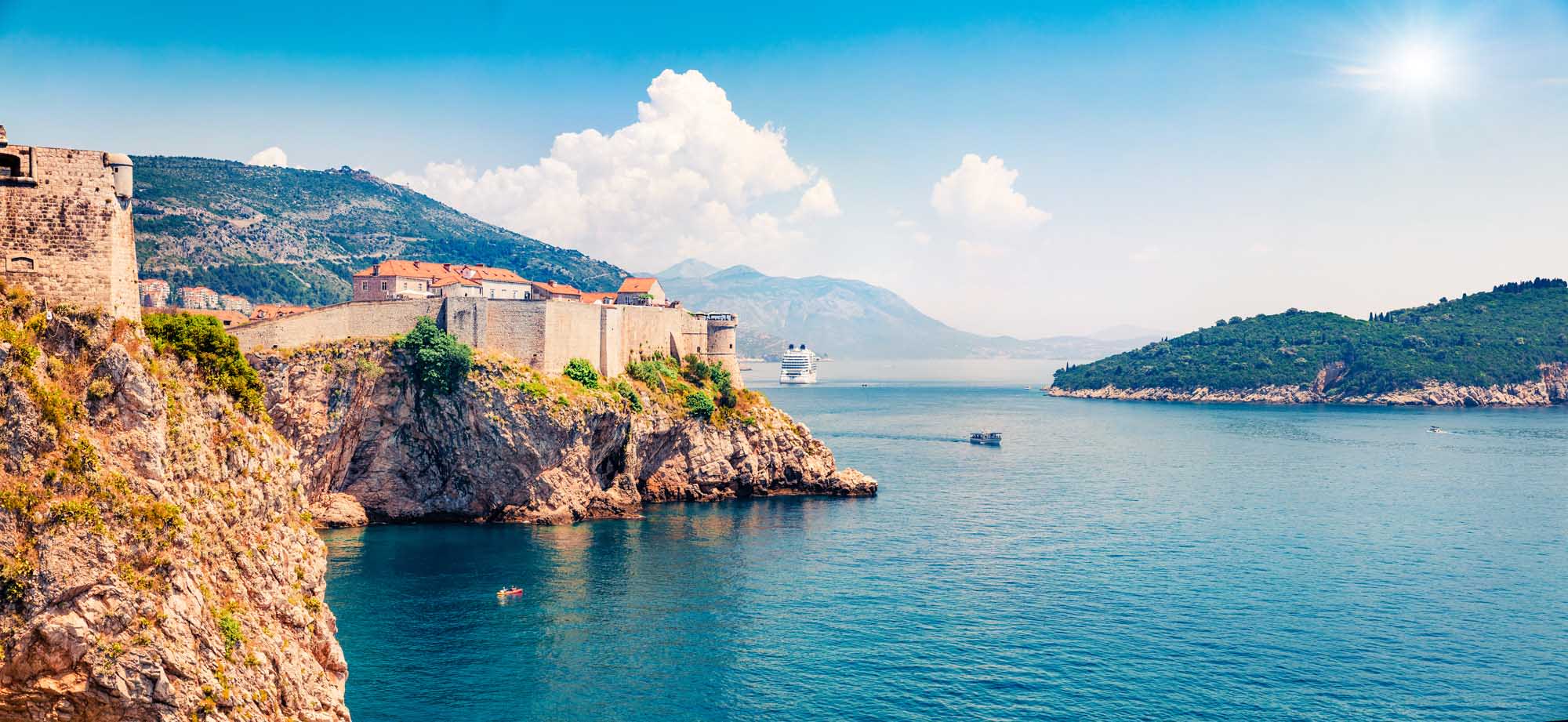 A panoramic view of the iconic Fort Bokar fortress perched on rocky cliffs in Dubrovnik, Croatia, overlooking the turquoise Adriatic Sea. A large cruise ship is visible in the harbor, with lush green islands and mountains in the background under a bright blue sky with white clouds.