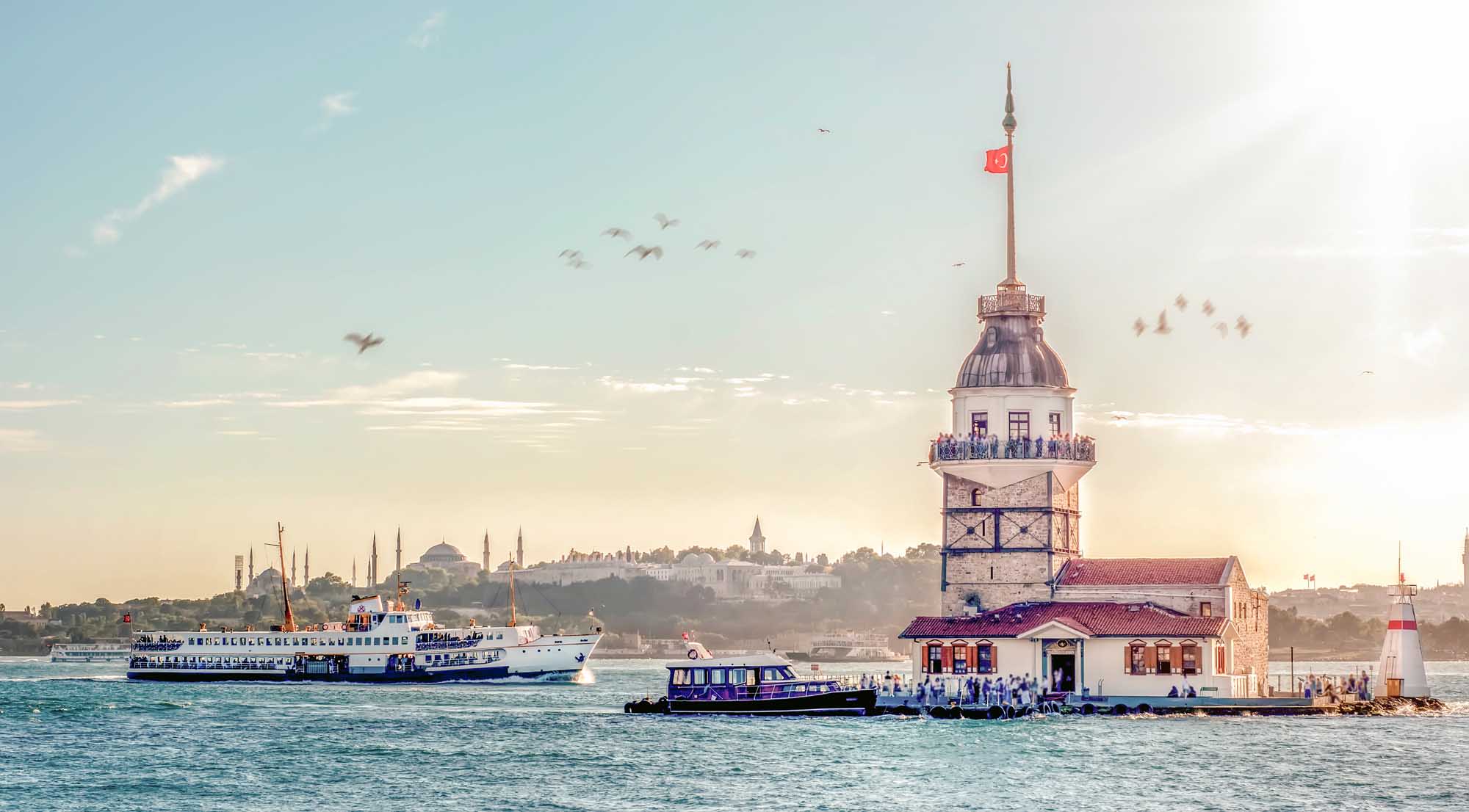 A scenic view of Istanbul's iconic Maiden's Tower (Kiz Kulesi) in the Bosphorus Strait, with a blue and white passenger ferry approaching the tower and the cityscape of Istanbul visible in the background under a clear sky. The historic lighthouse-style tower features a domed top with Turkish flag, surrounded by water taxis and sightseeing vessels.
